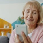 Idosa sorrindo enquanto usa o celular sozinha, sentada no sofá da sala