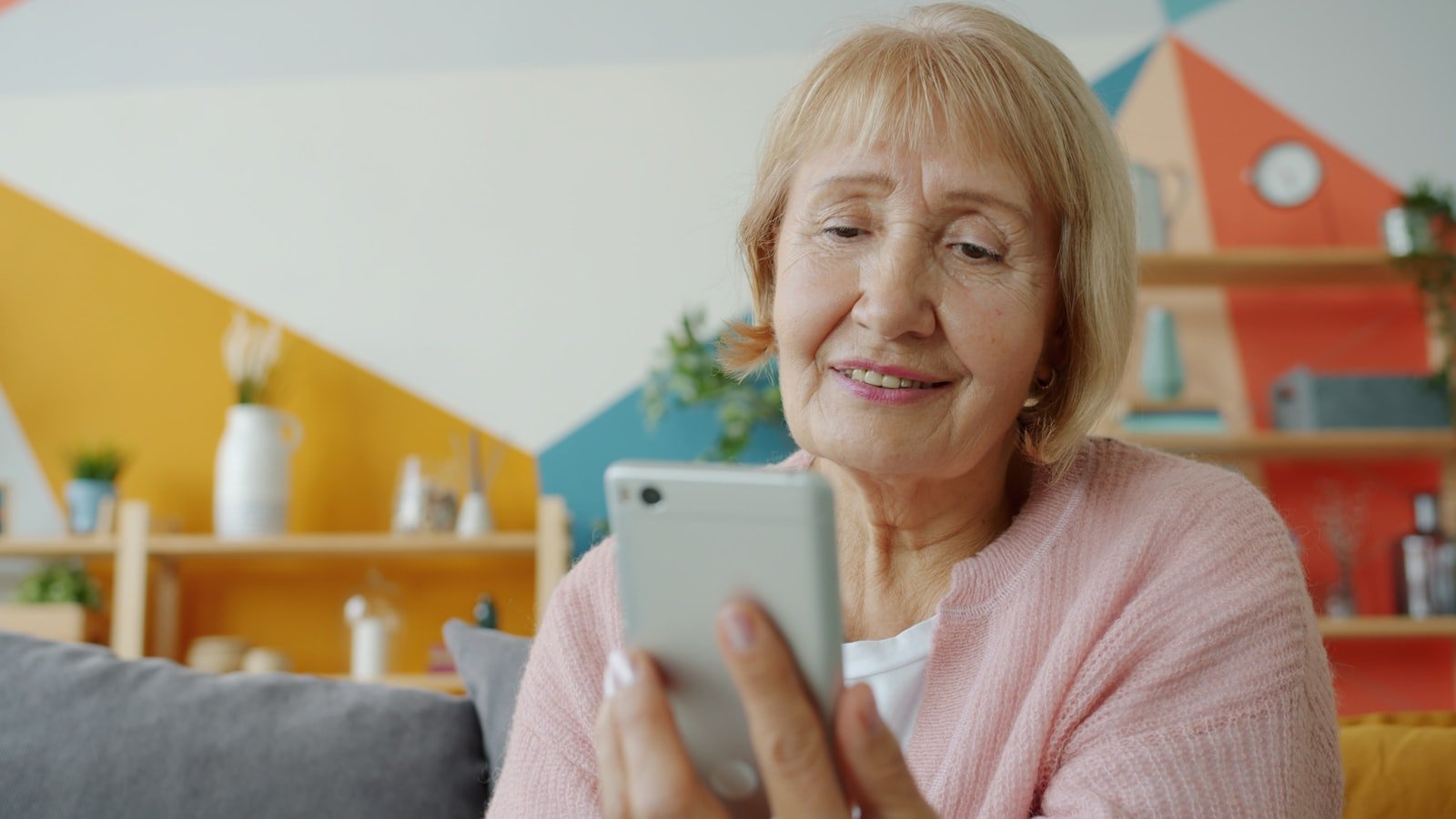 Idosa sorrindo enquanto usa o celular sozinha, sentada no sofá da sala