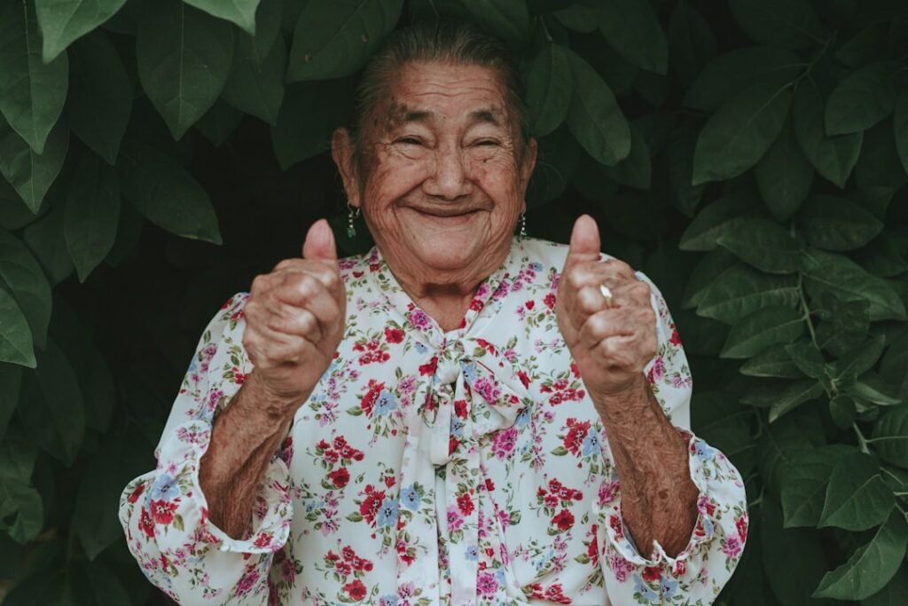 Idosa sorrindo com alegria genuína, fazendo sinal de positivo com as duas mãos, em jardim verde