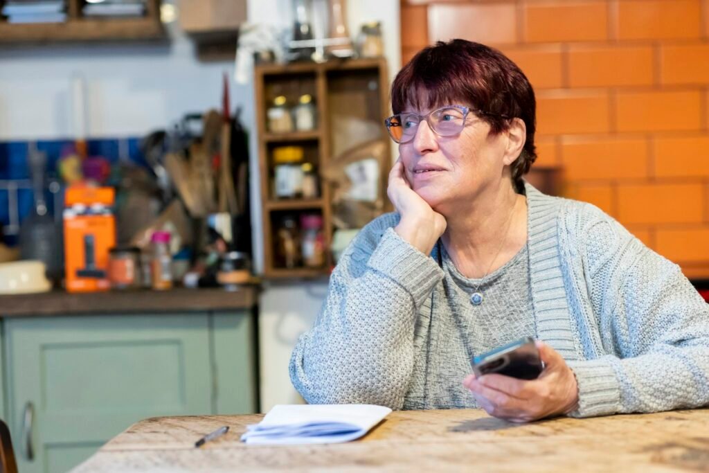 Mulher idosa pensativa apoiada na mesa da cozinha, segurando o celular
