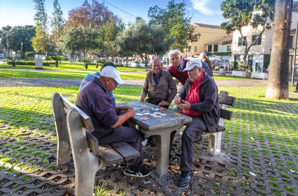 Grupo de cinco idosos reunido numa mesa de praça pública, jogando cartas ao ar livre num dia ensolarado