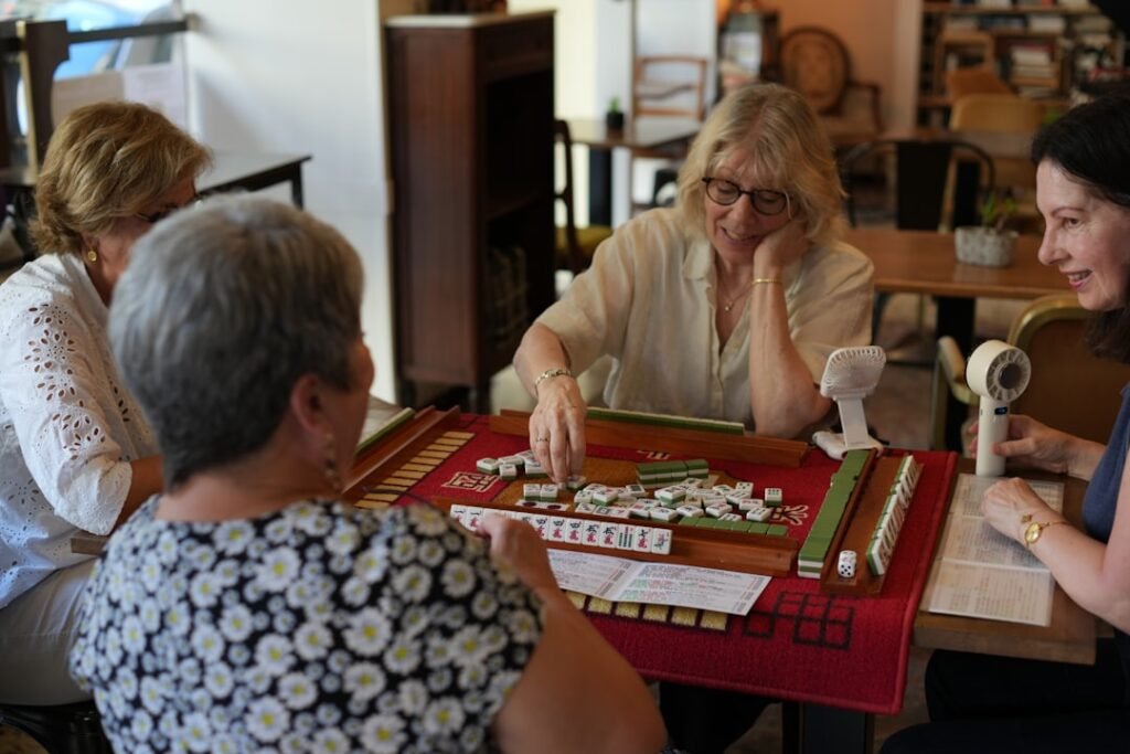 Grupo de quatro mulheres reunidas jogando mahjong numa mesa, sorrindo e conversando
