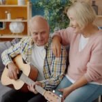 Casal de idosos sorrindo em casa enquanto o homem toca violão