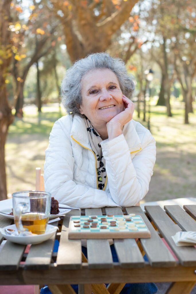 Idosa sorridente ao lado de um tabuleiro de damas, com café e um docinho sobre a mesa ao ar livre
