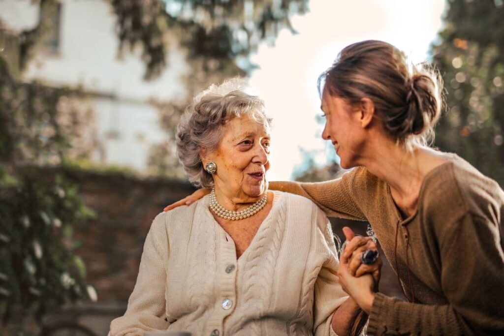 Idosa sorrindo enquanto é abraçada com carinho por uma cuidadora, em um jardim